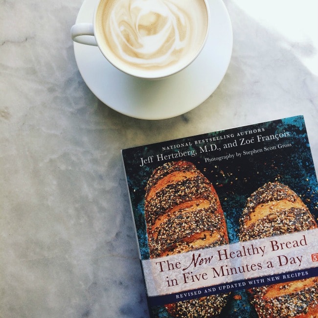 A cup of coffee on a table next to The New Healthy bread in Five Minutes a Day cookbook