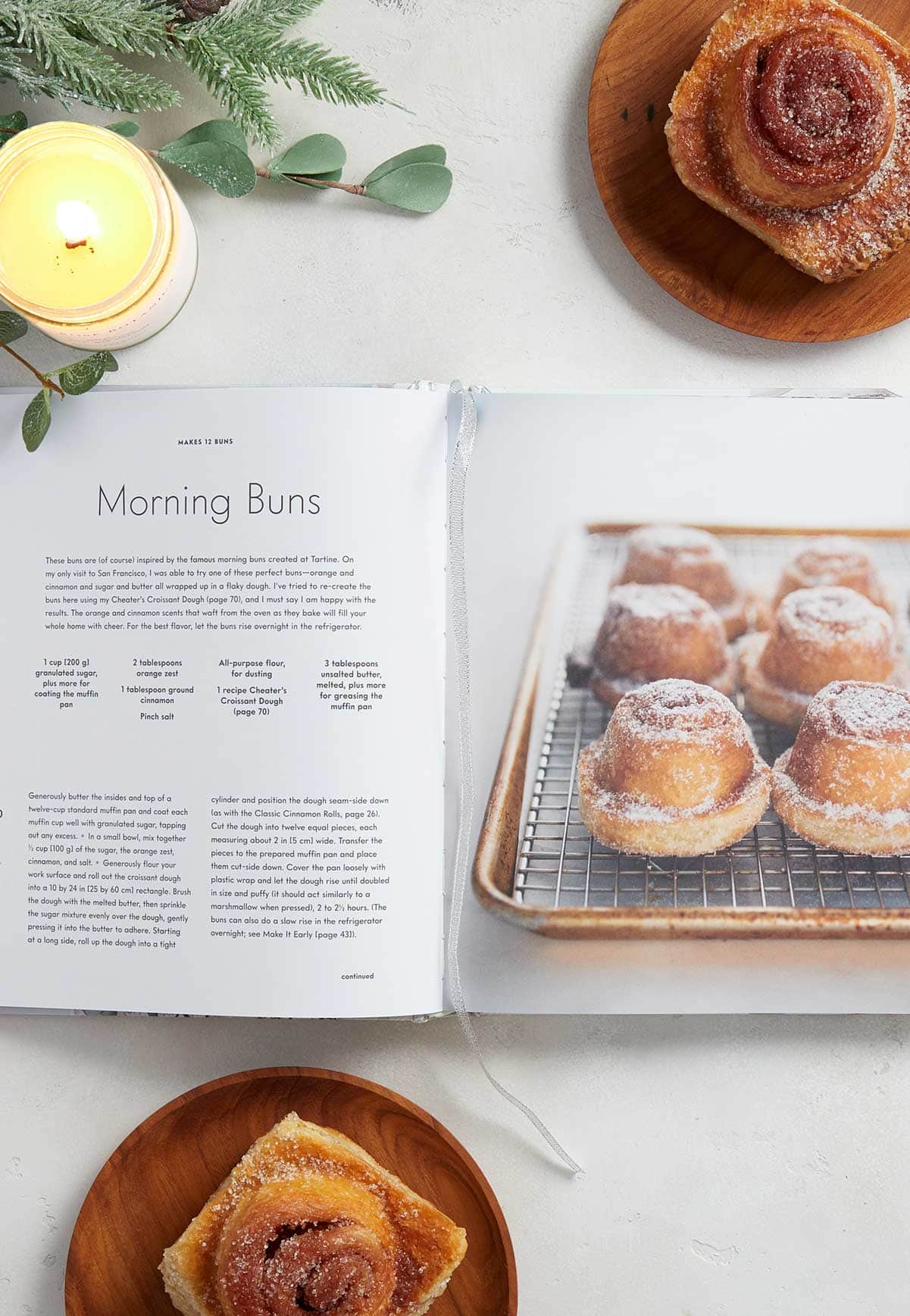 Open cookbook pages with a candle and morning buns beside it